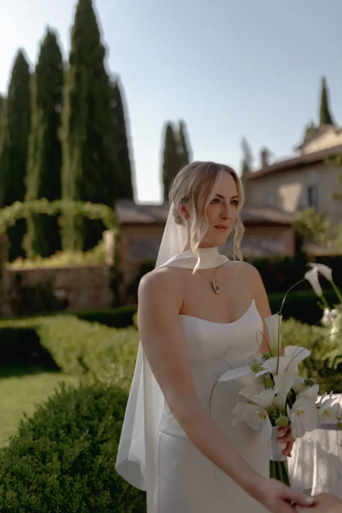 A bride in a white dress and veil holds a bouquet of white flowers, standing in a sunlit garden with tall trees and a building in the background.