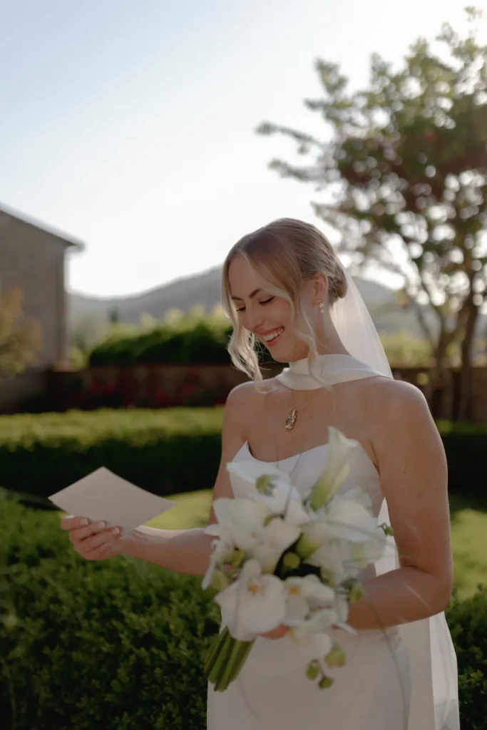 A bride in a white dress and veil smiles while reading a letter outdoors. She holds a bouquet of white flowers, with greenery and sunlight in the background.