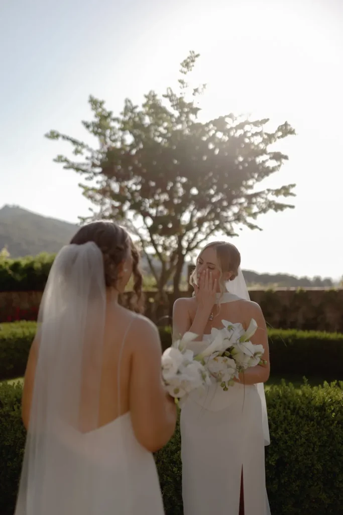 Two brides in white dresses and veils stand outdoors in a garden. One bride covers her mouth in surprise or joy, holding a bouquet, while the other faces her. Sunlight and a tree are visible in the background.