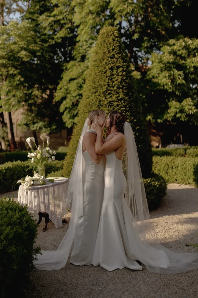 Two brides in white gowns and veils share a gentle, intimate moment in a sunlit garden, standing close together near a table with flowers, surrounded by greenery.