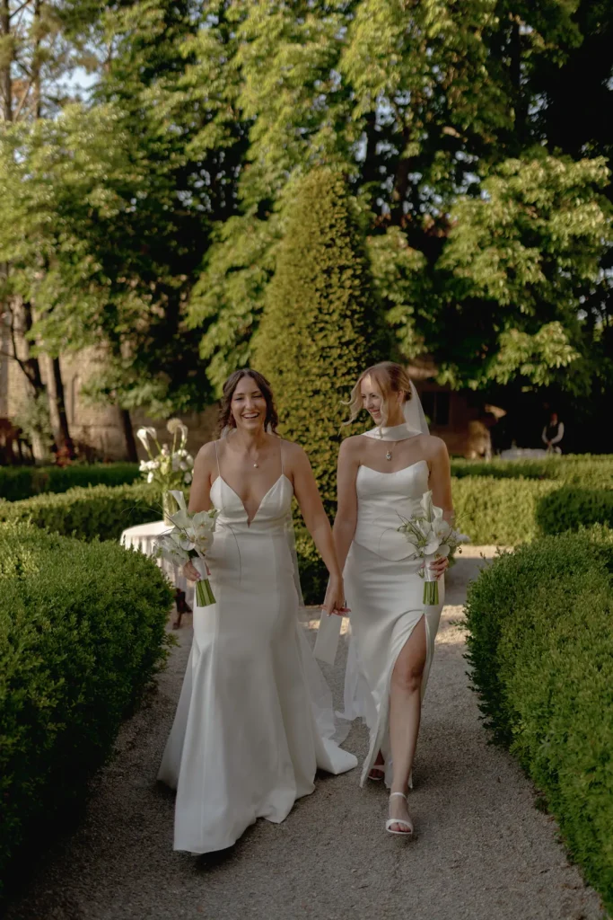 Two brides in white gowns walk hand in hand down a garden path, smiling at each other and holding bouquets, surrounded by greenery and sunlight.