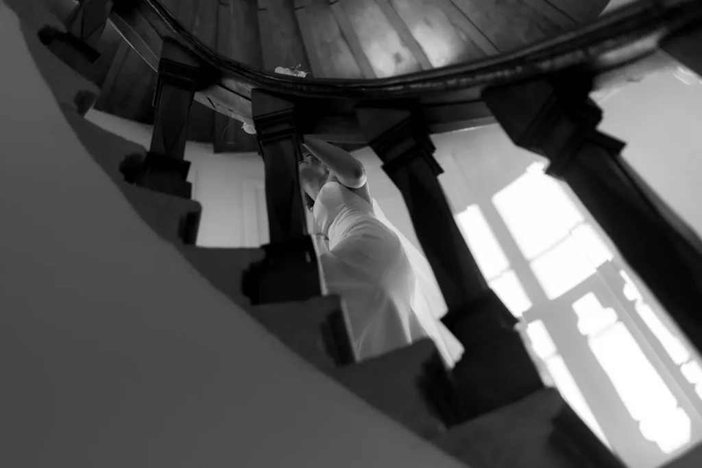 Black and white photo taken from below a spiral staircase, showing a person in a dress walking upward. Sunlight streams through tall windows, casting shadows on the stairs and wall.