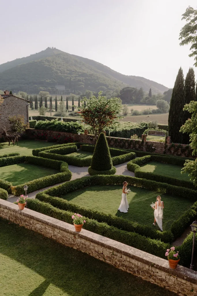 Two women in white dresses walk through a manicured garden with geometric hedges, surrounded by stone walls, potted plants, trees, and rolling hills in the background under a clear sky.