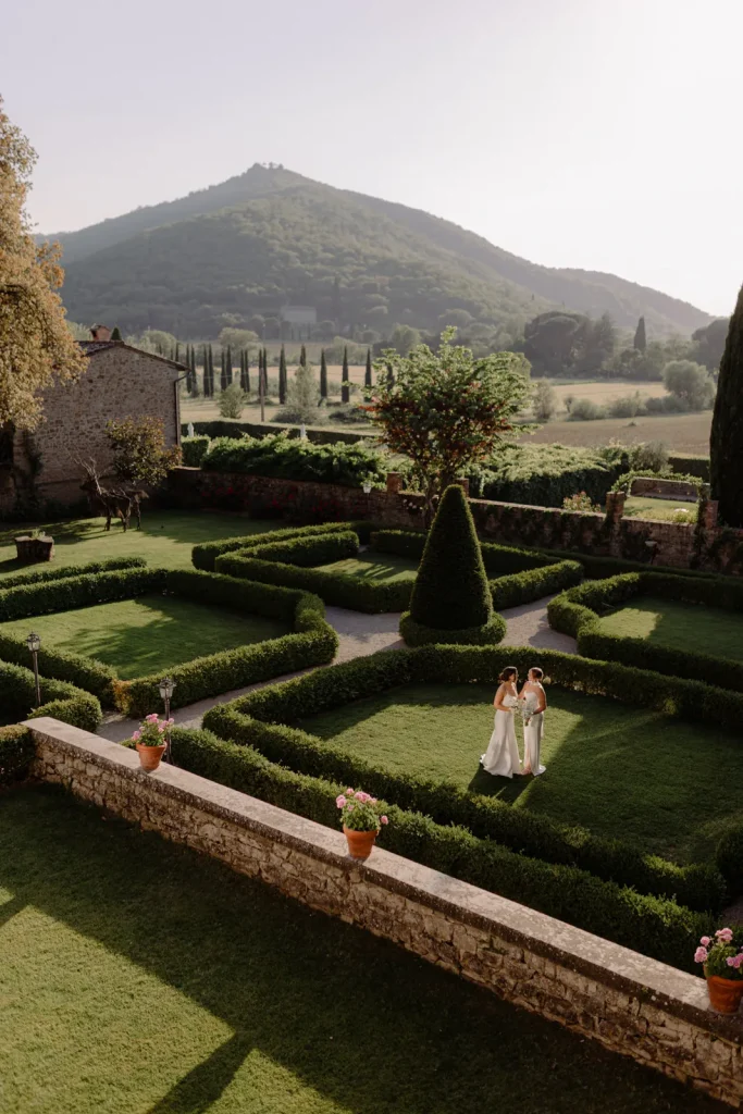 Two people in formal attire stand in a manicured garden with hedges, stone walls, and potted flowers, with distant hills and trees in the background under soft sunlight.