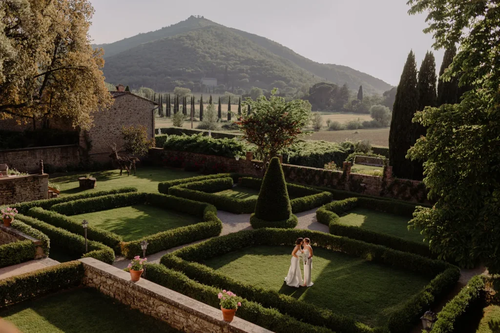 A couple in wedding attire stands in a manicured garden, surrounded by hedges and trees, with mountains and countryside visible in the background under soft sunlight.