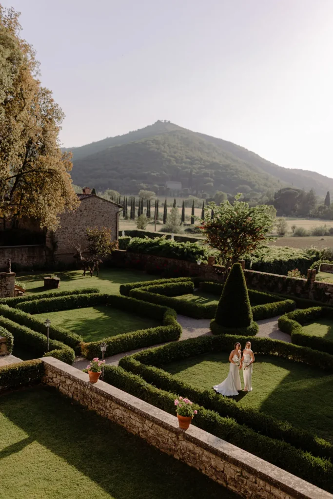 Two women in white dresses stand in a formal garden with manicured hedges and potted flowers, set against rolling hills and a distant mountain under a clear sky.