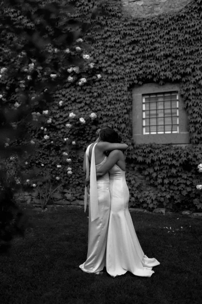 Two people in elegant dresses embrace in front of a window on a vine-covered wall. The photo is black and white, capturing a tender moment in an outdoor, garden-like setting.