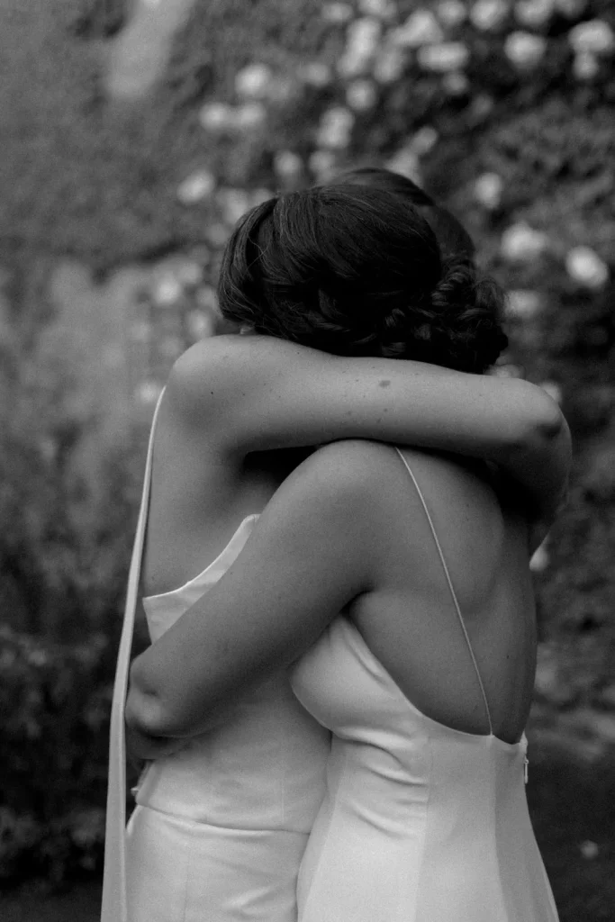 Two women in sleeveless dresses embrace each other closely outdoors. The moment is intimate and appears emotional. The image is in black and white, with a blurred background of foliage.