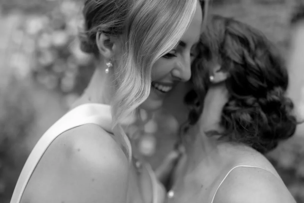 Two women, dressed formally, smile closely together in an affectionate moment. The black-and-white image captures joy and intimacy, with one woman’s face fully visible and the other turned away.
