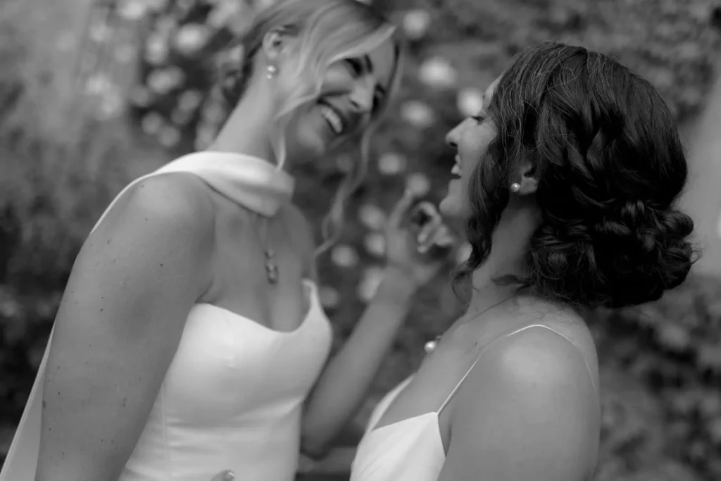 Two women in white dresses smile joyfully at each other, sharing a happy moment outdoors. The photo is black and white, with soft focus on their faces and blurred floral background.