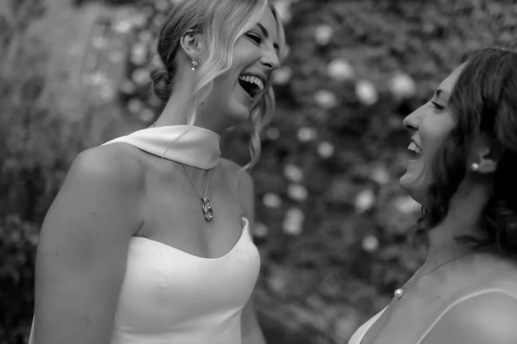 Two women in white dresses laughing joyfully together outdoors, with one wearing a scarf around her neck. The background is blurred with what appear to be flowers or foliage. Black and white photo.