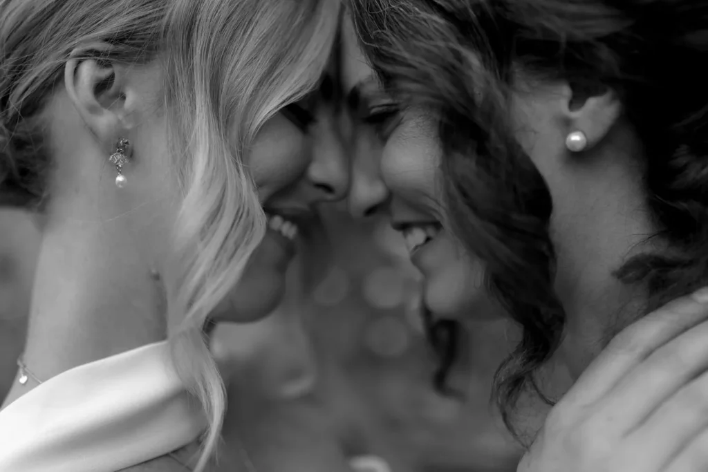 Two women smiling with closed eyes, foreheads touching closely in an affectionate, intimate moment. One has wavy hair and pearl earrings; the other has curled hair and also wears a pearl earring. The image is in black and white.