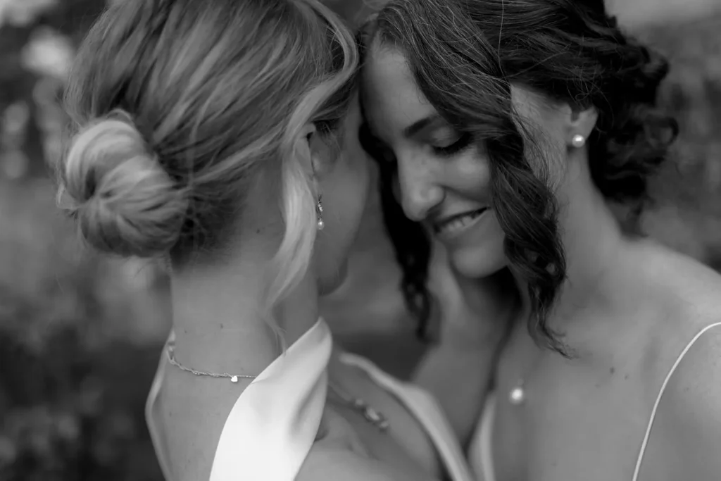 Two women in wedding dresses stand close together, touching foreheads and smiling softly. The photo is in black and white, highlighting their intimate and joyful moment.
