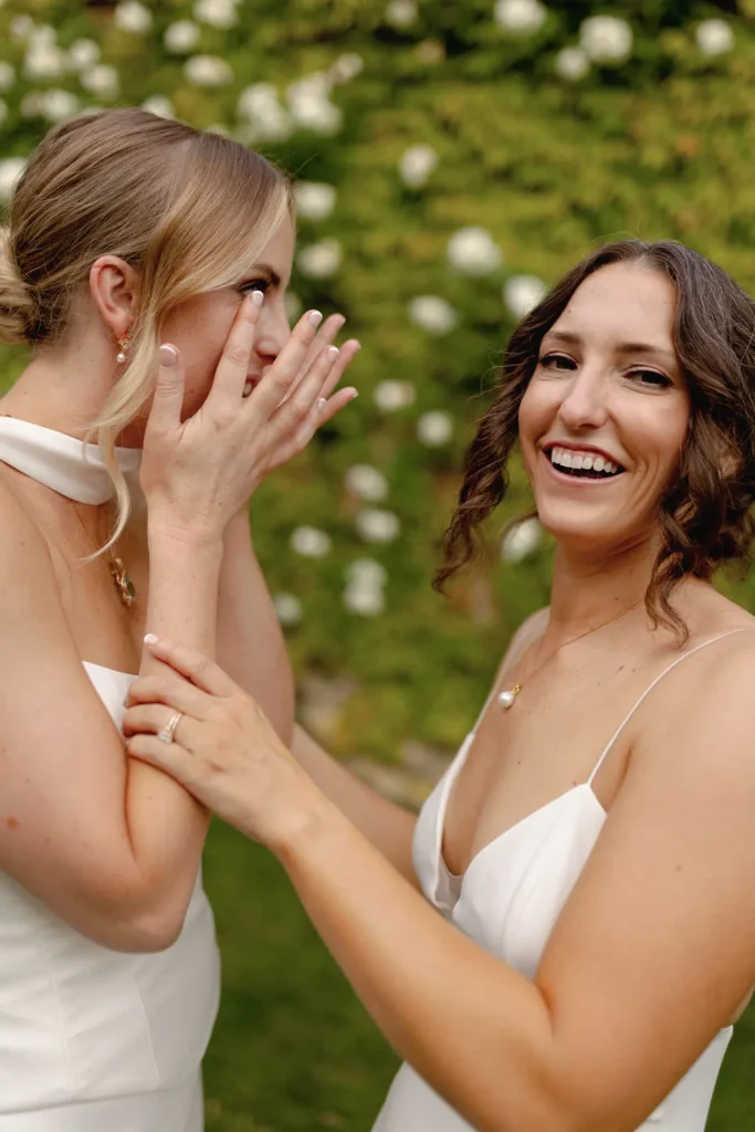 Two women in white dresses stand outdoors. One covers her mouth emotionally while the other holds her arm and smiles joyfully. Green foliage and white flowers are visible in the background.