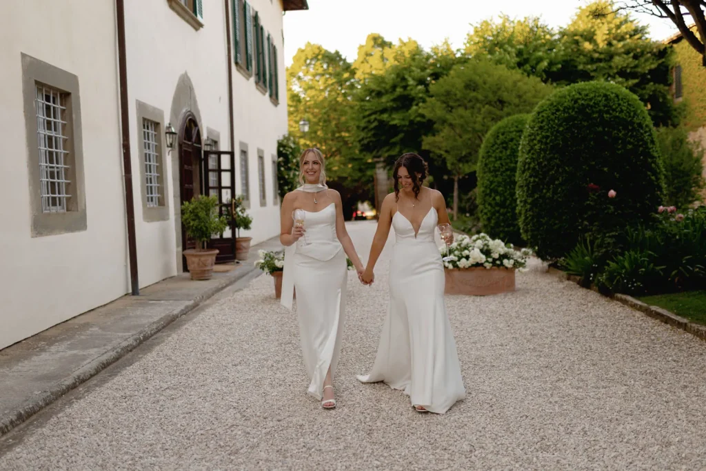 Two women in white wedding dresses walk hand in hand on a gravel path outside a large building with greenery and flowers, smiling and enjoying their moment together.