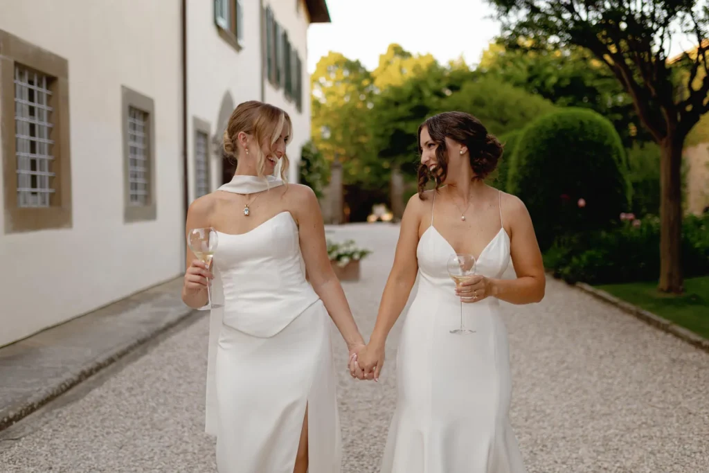Two women in elegant white dresses hold hands and smile at each other while walking outside on a gravel path, each holding a glass of wine, surrounded by greenery and buildings.