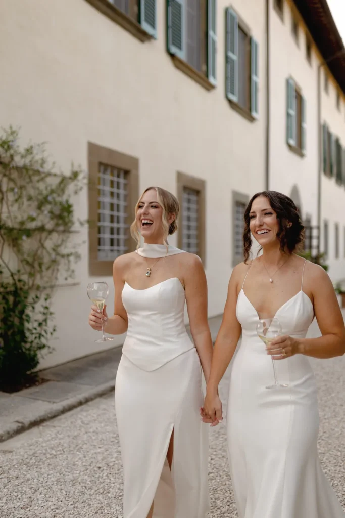 Two women in elegant white dresses hold hands and smile while walking outside a building, each carrying a glass of wine.