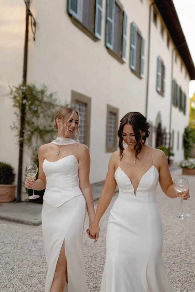 Two women in elegant white dresses hold hands and smile at each other, each holding a glass of white wine, walking outside a building with green shutters and arched doorways.