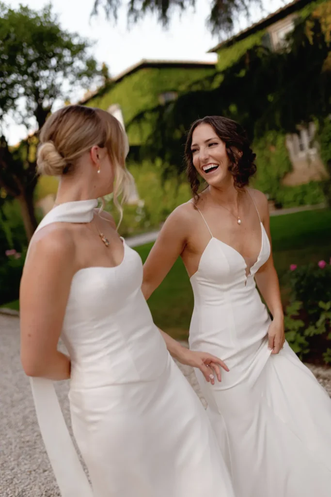 Two women in elegant white dresses smile and walk outdoors on a gravel path, surrounded by greenery and trees, on what appears to be a celebratory occasion.