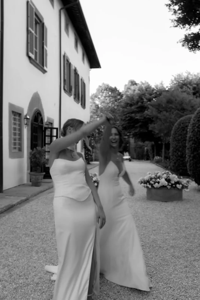 Two women in white dresses laugh and dance together outside a large building, surrounded by greenery and flowers, in a joyful, candid black-and-white moment.