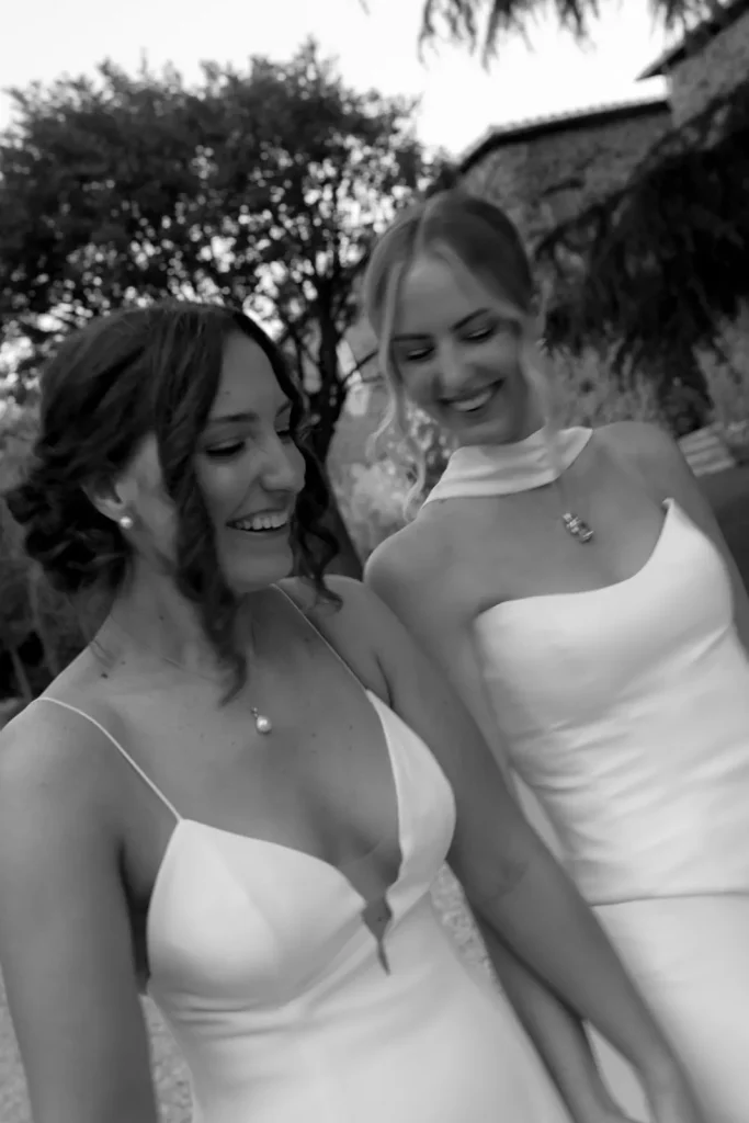 Two women in white dresses, smiling and holding hands outdoors. The image is black and white, with trees and a stone building in the background.