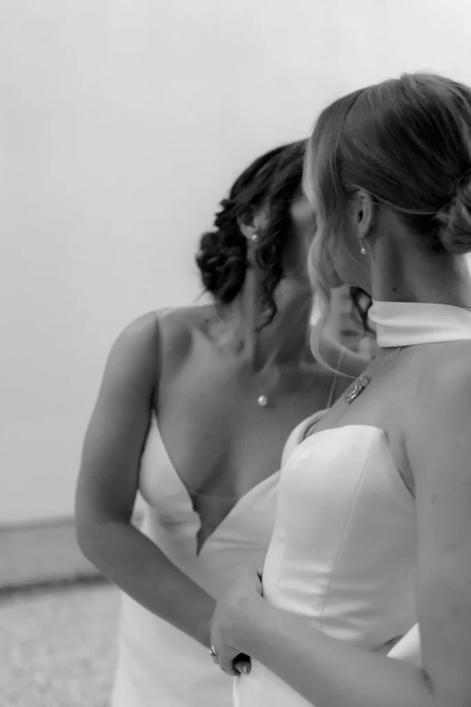 Two women in white dresses face each other closely, with their hair styled in updos. The photo is in black and white, capturing an intimate moment between them.
