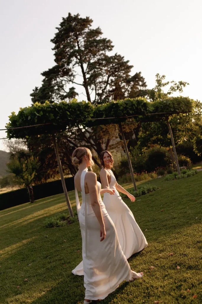 Two women in white wedding dresses walk together on a grassy lawn at sunset, surrounded by trees and greenery.