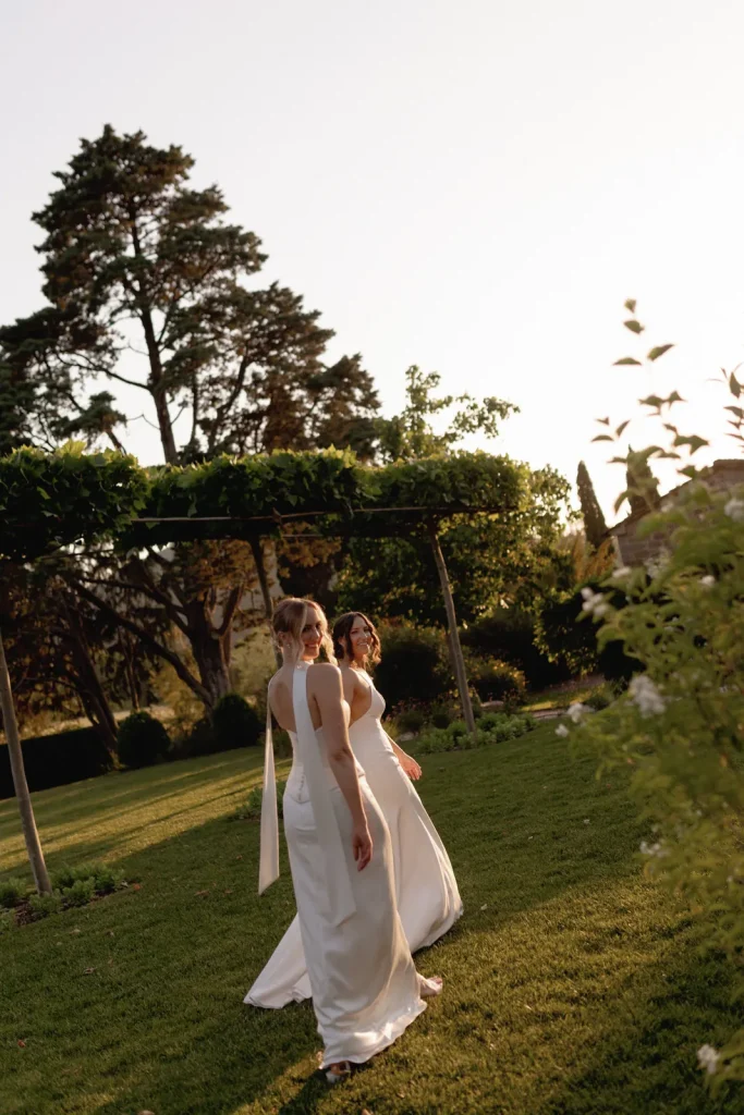 Two women in white dresses walk side by side across a sunlit garden, greenery and tall trees in the background, as they smile and glance back over their shoulders.