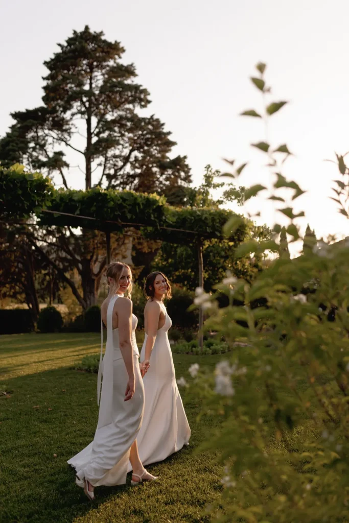 Two women in white dresses hold hands and walk through a sunlit garden, looking back toward the camera. Green foliage and tall trees surround them, creating a peaceful, romantic outdoor setting.