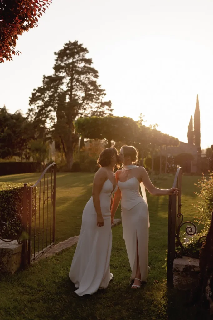 Two women in white dresses stand at a garden gate, sharing a tender moment at sunset, with sunlight casting a warm glow over the lush, green landscape and trees in the background.