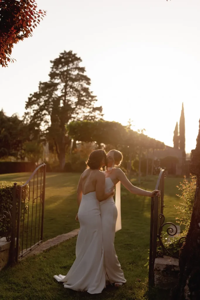 Two women in white dresses stand close, embracing and kissing outdoors by an open garden gate at sunset, surrounded by lush greenery and tall trees.