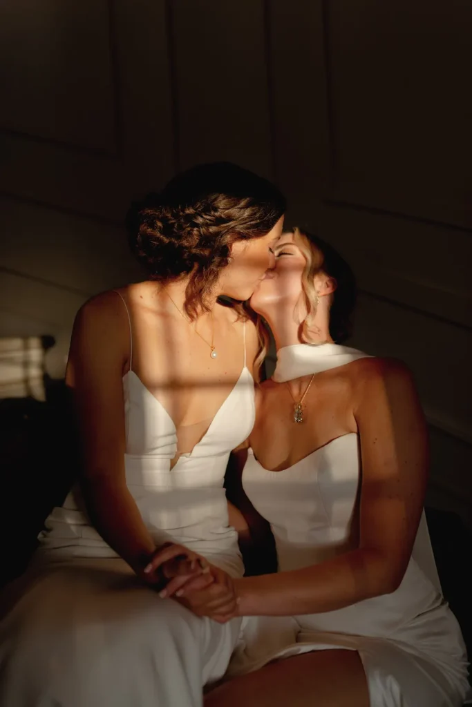 Two women in white dresses share a kiss while holding hands, softly lit by sunlight streaming through blinds, creating warm stripes of light and shadow across their faces and bodies.