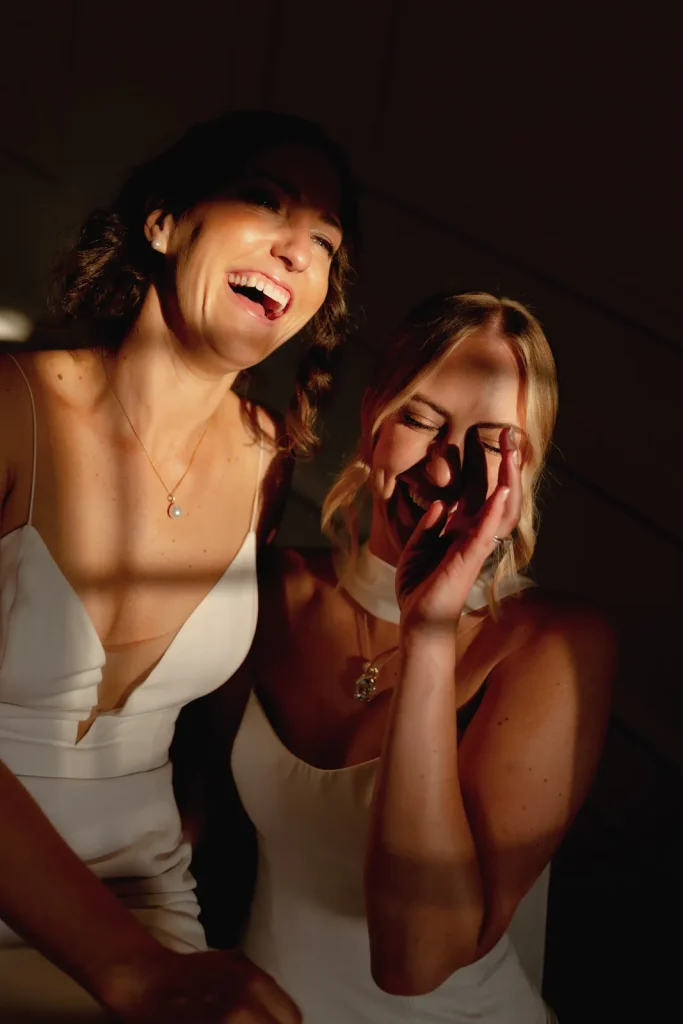 Two women in white dresses laugh joyfully together in warm sunlight, casting soft shadows across their faces. One woman covers part of her face with her hand, and both appear happy and relaxed, sharing a candid, joyful moment.