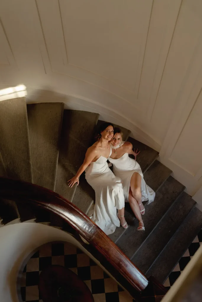 Two women in white dresses sit together on a curved staircase, smiling up at the camera. Sunlight softly illuminates part of the scene, creating a warm and joyful atmosphere.
