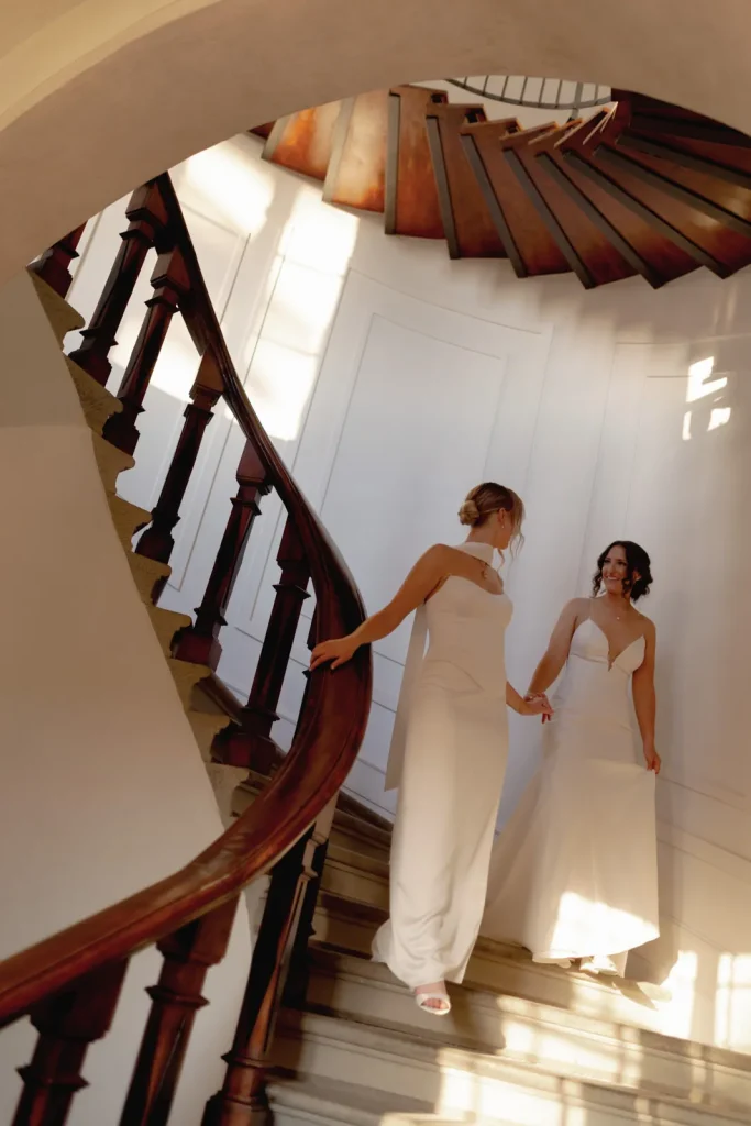 Two women in white dresses hold hands while descending a spiral staircase, sunlight streaming through windows and creating shadows on the walls.