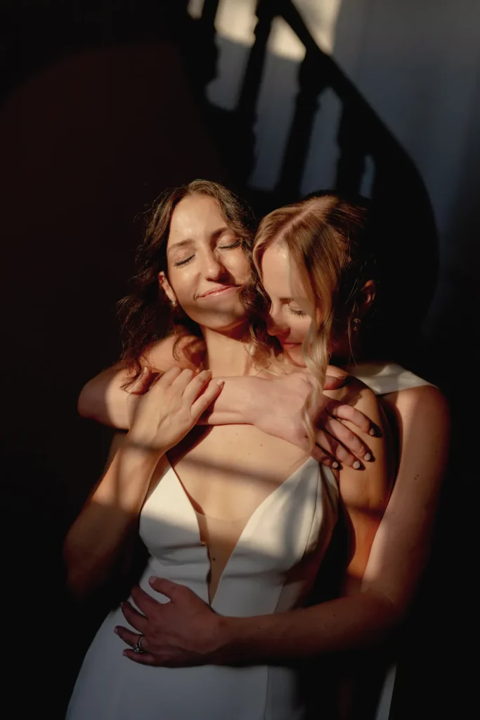 Two women in white dresses embrace lovingly in warm sunlight indoors, with one standing behind the other, arms wrapped around her partner, both smiling peacefully. Shadows from a nearby staircase fall across them.