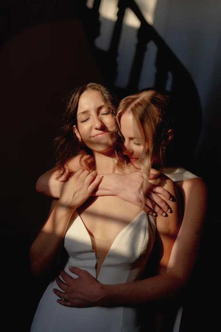 Same-Sex Wedding in Italy at Villa di Piazzano 1 Two women in white dresses embrace lovingly in warm sunlight indoors, with one standing behind the other, arms wrapped around her partner, both smiling peacefully. Shadows from a nearby staircase fall across them.