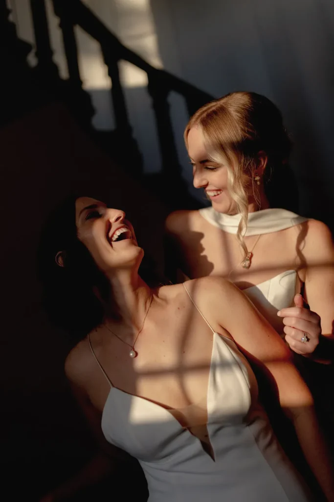 Two women in white dresses smile and embrace in soft, dramatic sunlight indoors, with shadows and a staircase in the background.