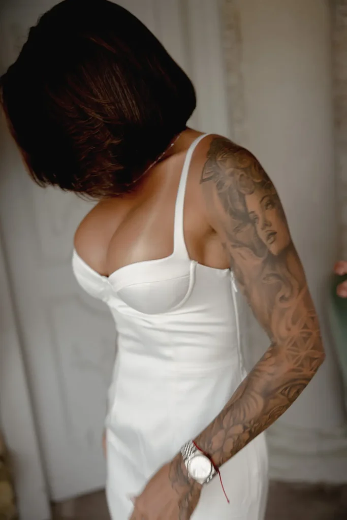 Luxury Couple Shoot in Taormina | Italian Glamour 43 Woman in a white dress with brown bobbed hair. Her arm has an intricate tattoo featuring a womans face and patterns. She is looking down and away, inside a softly lit room.