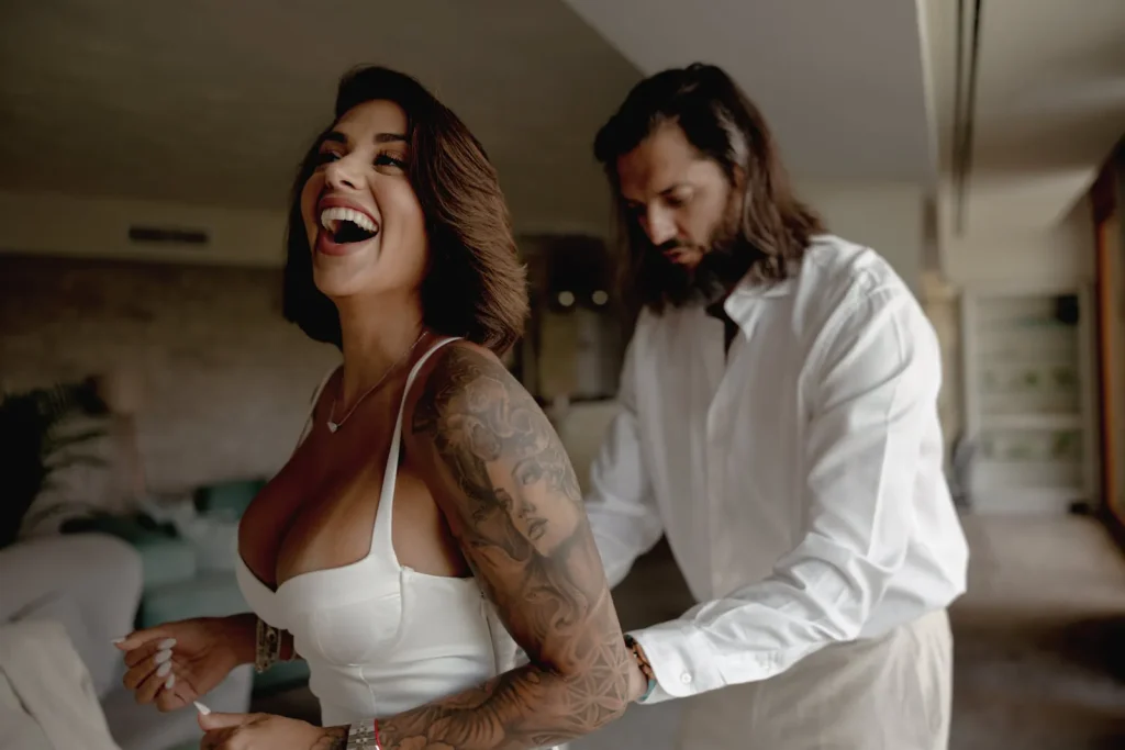 Luxury Couple Shoot in Taormina | Italian Glamour 67 A woman in a white dress laughs as a man in a white shirt helps zip her dress from behind in a bright living room.