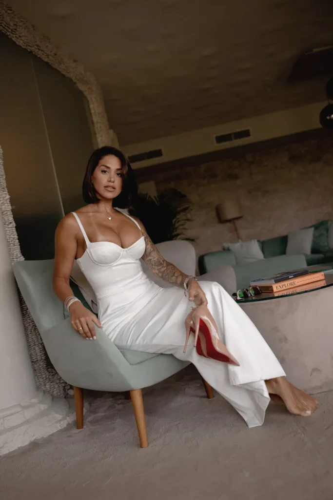 Luxury Couple Shoot in Taormina | Italian Glamour 52 Woman in a white dress sits on a light green chair, holding red high heels. She has a tattoo on her left arm, short dark hair, and jewelry. There are books on a table and a neutral-toned room in the background.