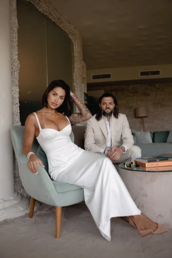 Luxury Couple Shoot in Taormina | Italian Glamour 64 A woman in a white dress sits on a chair, while a man in a light suit sits on a couch behind her. They are in a modern living room with books on the table.
