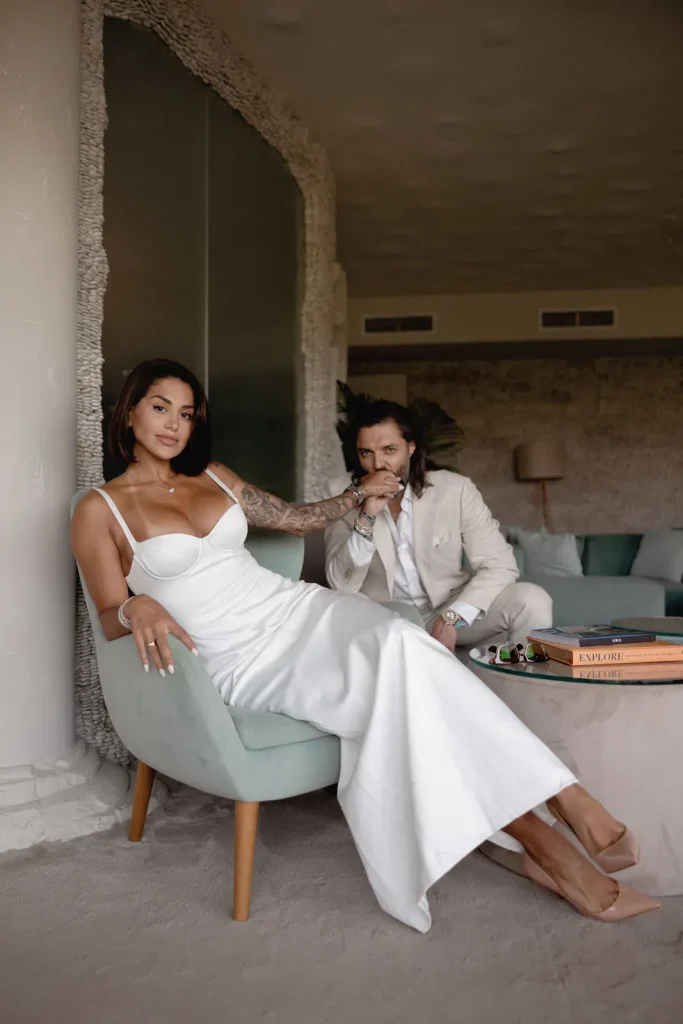 Luxury Couple Shoot in Taormina | Italian Glamour 18 A woman in a white dress sits on a chair, with a man in a light suit kneeling beside her, gently kissing her hand. They are in a stylish, modern room with a coffee table holding books.