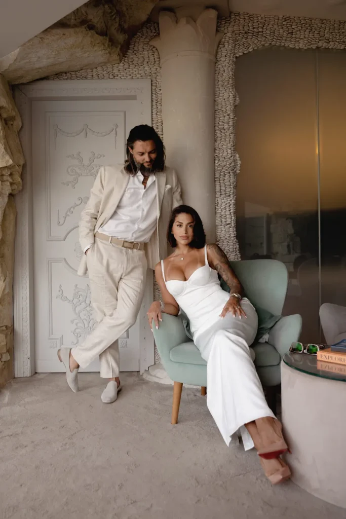 Luxury Couple Shoot in Taormina | Italian Glamour 46 A man in a beige suit stands next to a woman in a white dress seated in a light green chair. They pose inside a stylish room with ornate walls and a round table holding books and sunglasses.