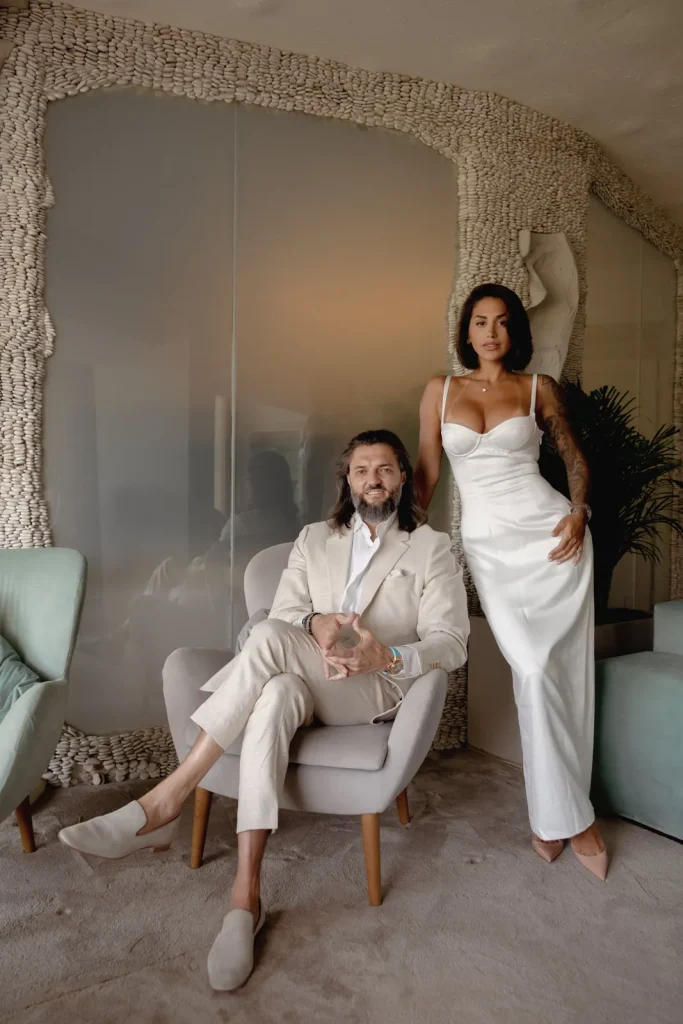 Luxury Couple Shoot in Taormina | Italian Glamour 60 A man in a beige suit sits in an armchair while a woman in a white dress stands beside him in a stylish, modern room with textured walls and soft furnishings.