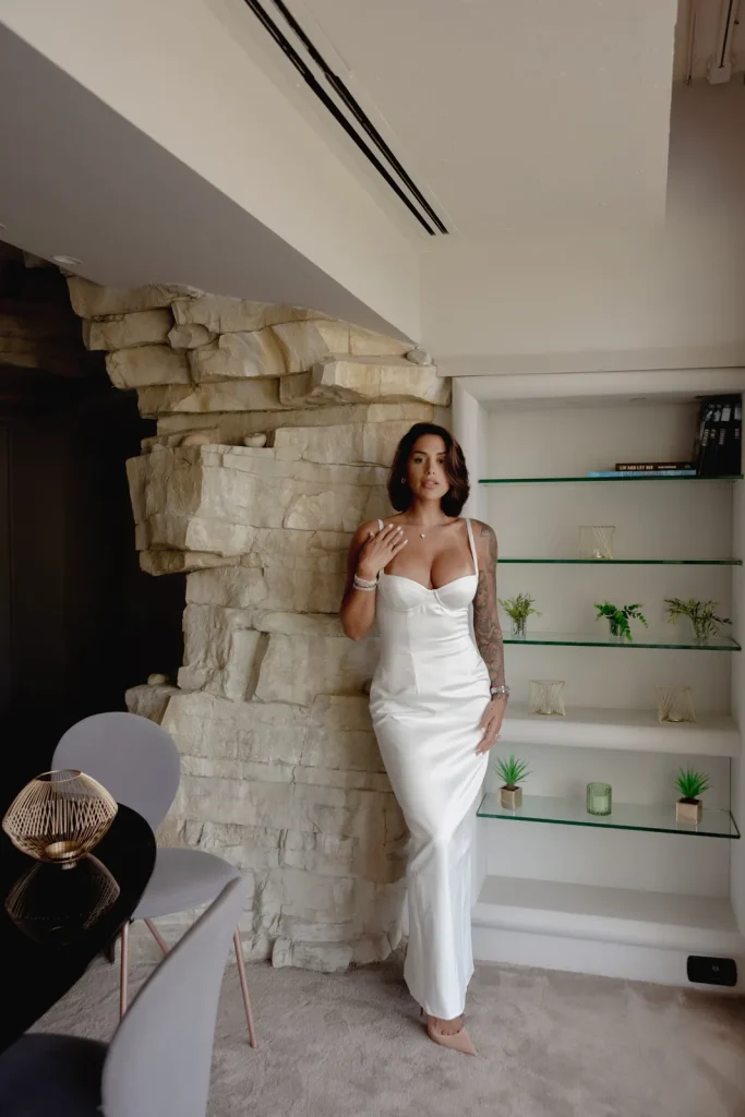Luxury Couple Shoot in Taormina | Italian Glamour 49 A woman in a white dress stands by a stone wall and glass shelves with small plants and decor items in a modern room.