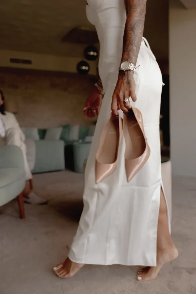 Luxury Couple Shoot in Taormina | Italian Glamour 15 A woman in a white dress walks barefoot indoors, holding nude high heels in one hand. Her arm features tattoos and a watch. Another person sits blurred in the background.