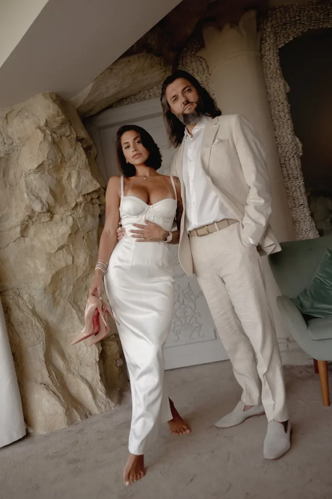 Luxury Couple Shoot in Taormina | Italian Glamour 66 A woman in a white satin dress and a man in a beige suit stand together indoors, next to a rock wall and a green chair. The woman holds pink heels and both look at the camera.