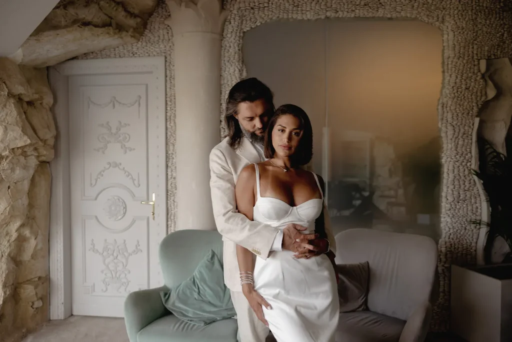 Luxury Couple Shoot in Taormina | Italian Glamour 19 A man in a light suit stands behind a woman in a white dress, holding her waist. They pose in a stylish room with ornate decor, a white door, and an armchair. Both look serious and elegant.