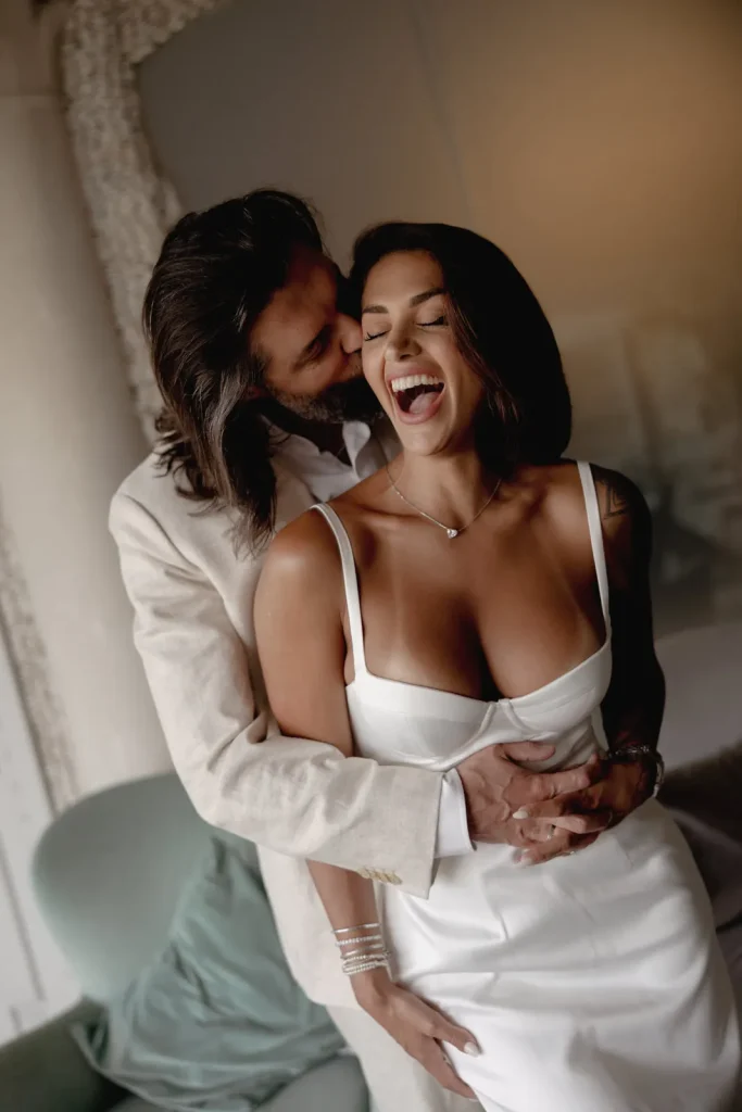 Luxury Couple Shoot in Taormina | Italian Glamour 30 A man in a light suit hugs and kisses a laughing woman in a white dress from behind. Both appear joyful and affectionate in a softly lit room.